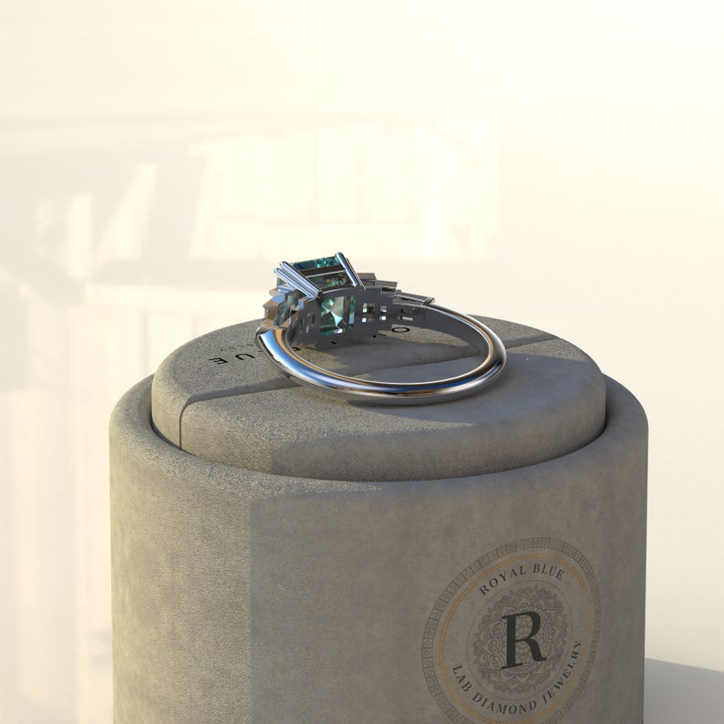 May include: A silver ring with a large emerald-cut green gemstone and smaller accent stones. The ring is sitting on a gray box with the text "Royal Blue Lab Diamond Jewelry" and a large "R" in the center.