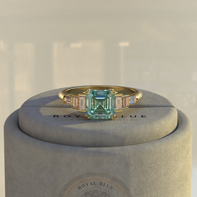 May include: A gold ring with a square-shaped green gemstone surrounded by smaller clear gemstones. The ring is in a box with the text "ROYAL BLUE LAB DIAMOND JEWELRY".