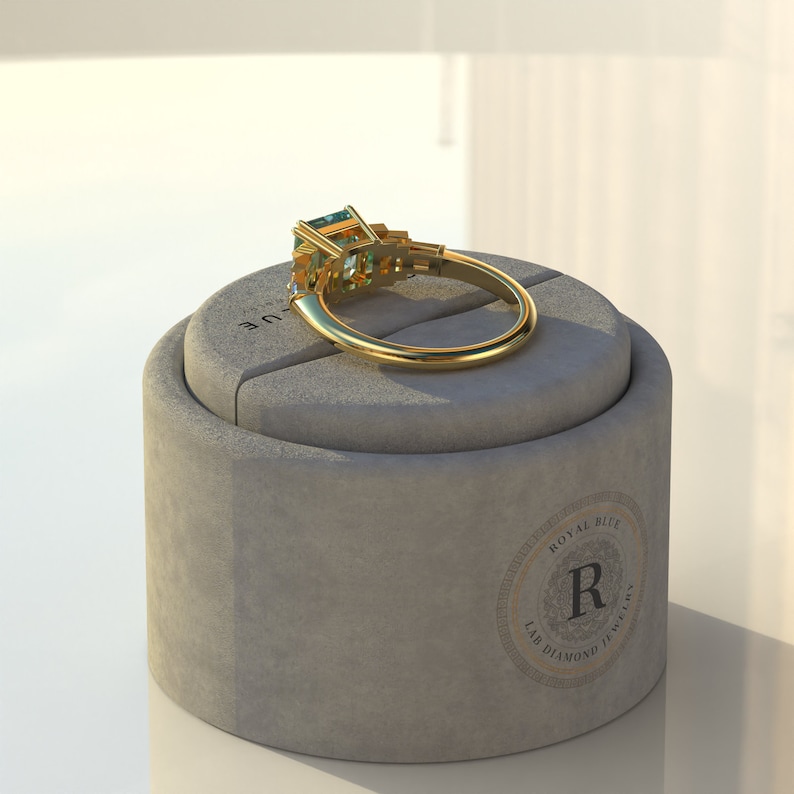 May include: A gold ring with a large emerald-cut green gemstone surrounded by smaller diamonds. The ring is displayed on a gray ring box with the text "Royal Blue Lab Diamond Jewelry" printed on the side.