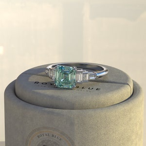 May include: A silver ring with a square emerald green gemstone and smaller baguette-shaped diamonds on the band. The ring is displayed in a gray box with the text "ROYAL BLUE LAB DIAMOND JEWELRY".