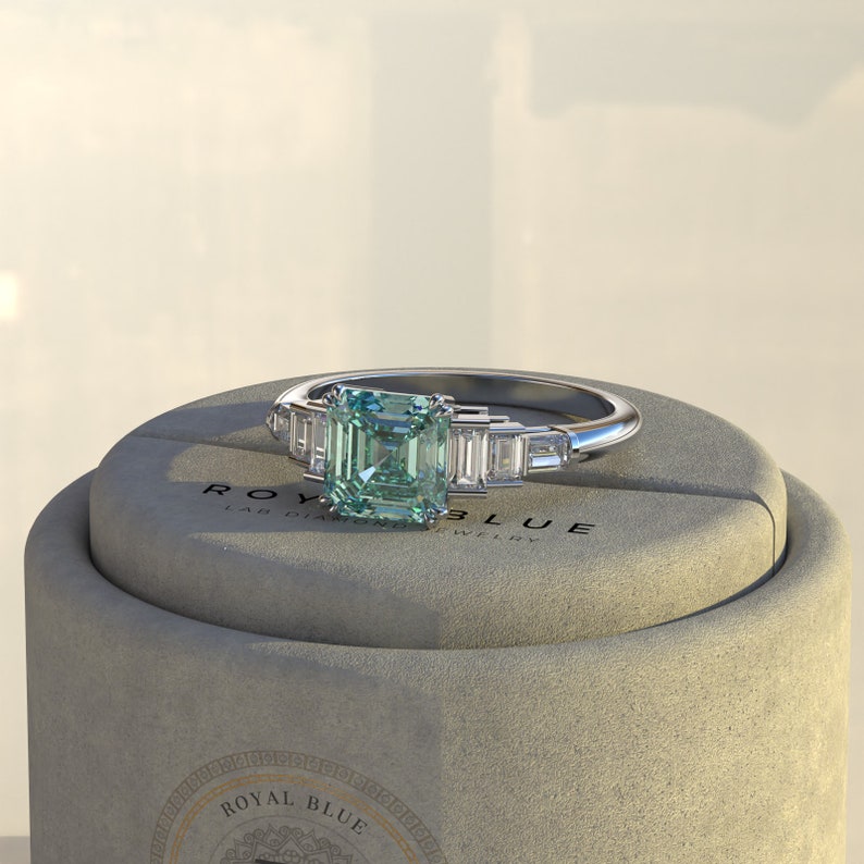 May include: A silver ring with a square emerald green gemstone and smaller baguette-shaped diamonds on the band. The ring is displayed in a gray box with the text "ROYAL BLUE LAB DIAMOND JEWELRY".