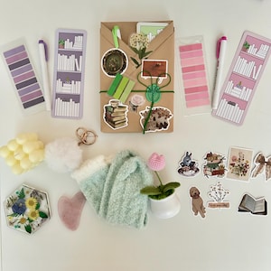 May include: A flat lay of a variety of items, including a brown paper package wrapped with green twine, a white pom-pom, a pair of fuzzy green socks, a small potted plant with a pink flower, a heart-shaped pink stone, a hexagonal resin tray with dried flowers, a set of yellow candles, a set of purple and pink sticky notes, a set of pink sticky notes, two white markers, two bookmarks with a bookshelf design, and a variety of stickers with book and floral themes. The stickers include a book stack, a teacup with a bunny, a floral bouquet, a book with the text "Love yourself", and a book with a floral design.