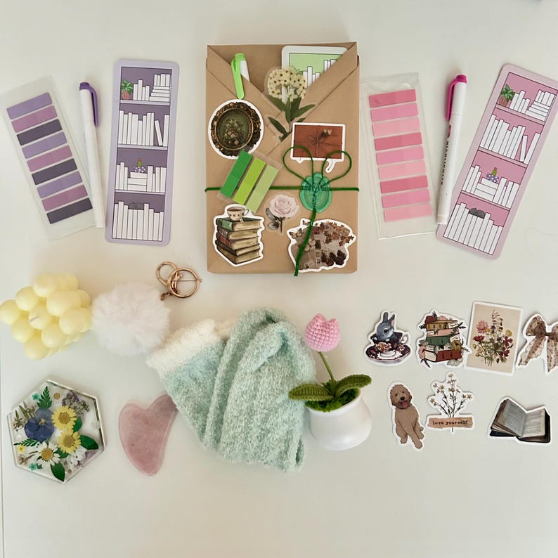 May include: A flat lay of a variety of items, including a brown paper package wrapped with green twine, a white pom-pom, a pair of fuzzy green socks, a small potted plant with a pink flower, a heart-shaped pink stone, a hexagonal resin tray with dried flowers, a set of yellow candles, a set of purple and pink sticky notes, a set of pink sticky notes, two white markers, two bookmarks with a bookshelf design, and a variety of stickers with book and floral themes. The stickers include a book stack, a teacup with a bunny, a floral bouquet, a book with the text "Love yourself", and a book with a floral design.