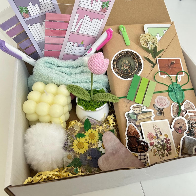 May include: A gift box filled with various items, including a crocheted flower, a small potted plant, a white fluffy pom-pom, a heart-shaped pink stone, a clear resin tray with pressed flowers, a candle, a small towel, and a brown envelope with stickers.