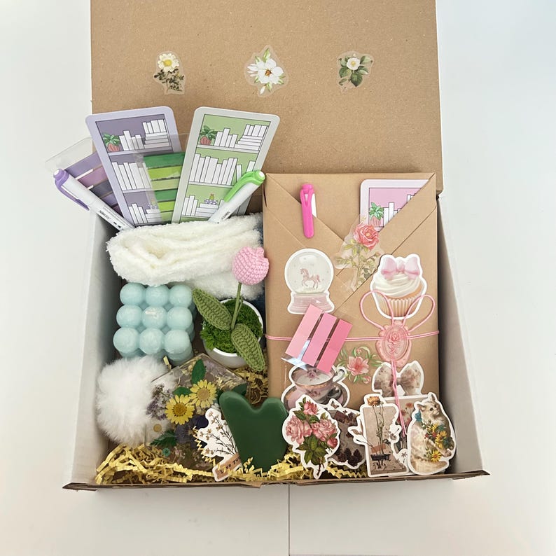 May include: A gift box filled with various items, including a white towel, a small potted plant, a blue bubble bath, a pink cupcake sticker, a snow globe sticker, and a brown envelope with a floral design. The box is filled with shredded paper.