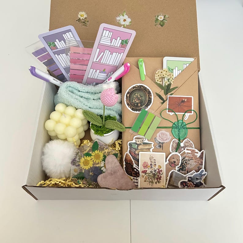 May include: A white box filled with a variety of items, including a small potted plant, a white fluffy ball, a pink heart, a yellow candle, and a variety of stickers and paper goods. The box is open and the items are visible.