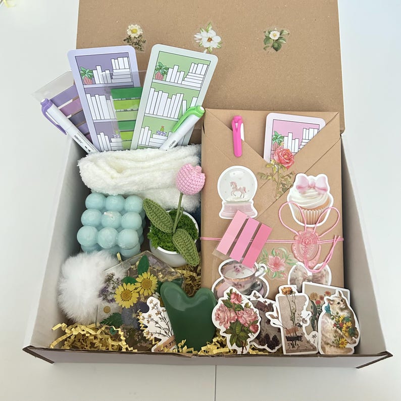 May include: A brown cardboard box filled with various items, including a white towel, a blue bubble bath, a green succulent, a pink flower, a white pom-pom, a green jade roller, a clear resin tray with dried flowers and a sunflower, a pink cupcake sticker, a pink snow globe sticker, a pink teacup sticker, a pink floral sticker, a cat sticker, and a floral sticker.