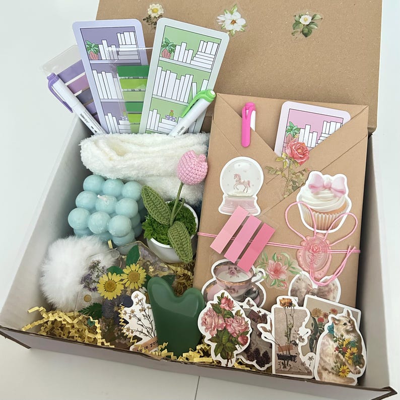 May include: A brown box filled with a variety of gifts, including a white towel, a blue bubble candle, a green crocheted plant, a pink flower, a snow globe, a cupcake sticker, a heart-shaped green stone, and a variety of floral stickers.