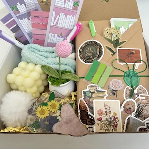 May include: A gift box filled with various items, including a crocheted flower, a candle, a small towel, a heart-shaped stone, a book-themed sticker sheet, and a brown envelope with stickers and a wax seal.