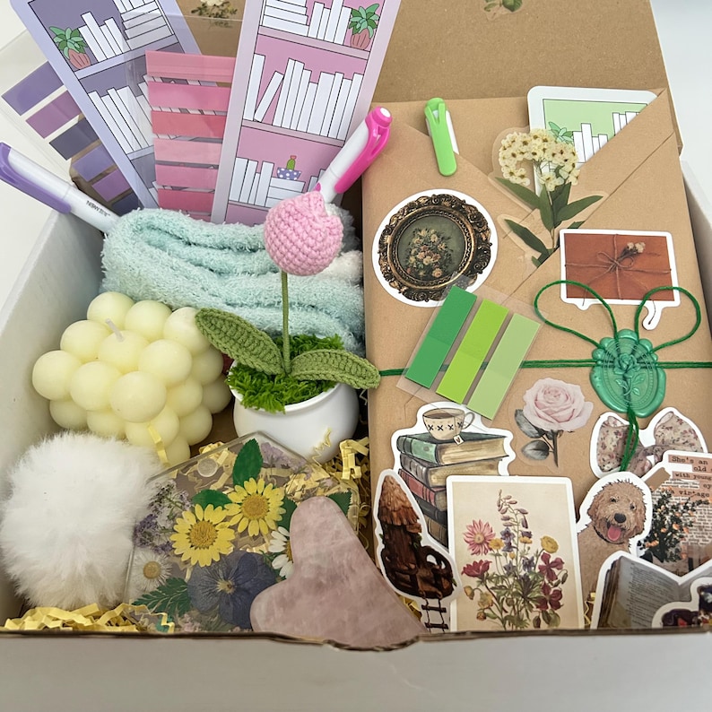 May include: A gift box filled with various items, including a crocheted flower, a candle, a small towel, a heart-shaped stone, a book-themed sticker sheet, and a brown envelope with stickers and a wax seal.