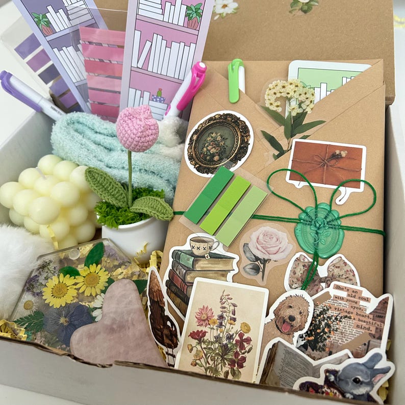 May include: A gift box filled with various items, including a brown envelope with stickers, a crocheted flower, a small potted plant, a heart-shaped resin piece with dried flowers, and a stack of books with stickers. The box is filled with shredded paper.