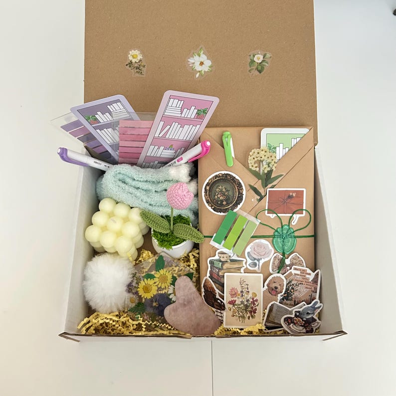 May include: A brown cardboard box filled with a variety of items, including a small potted plant, a white fluffy ball, a pink heart-shaped object, a stack of books, and various stickers. The box is open and the items are visible inside.
