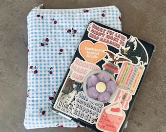 May include: A blue and white gingham Kindle sleeve with red cherry accents next to a black Kindle with stickers. The Kindle has a purple flower grip and the text "Sorry I'm late I didn't want to stop reading."
