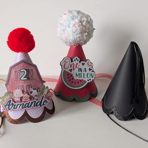 Könnte beinhalten: Drei Partyhüte. Einer ist rot mit einem roten Pompon, einer Scheunengrafik, der Zahl 2 und dem Namen "Armando". Ein weiterer ist rot mit einem weißen Pompon und einer "One in a Melon"-Grafik. Der dritte ist schwarz mit einem gewellten Rand.