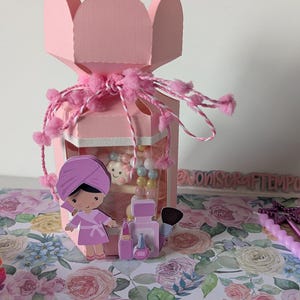 May include: A pink paper gift box with a spa theme, decorated with pink pom-poms and a ribbon. Inside, there's a spa-themed paper cutout of a girl in a robe, and beauty products. The box sits on a floral patterned surface.
