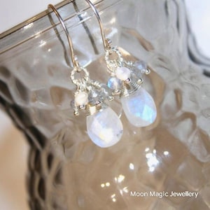 May include: Silver dangle earrings with moonstone and white pearl accents. The earrings feature a teardrop-shaped moonstone with a shimmering iridescent finish.