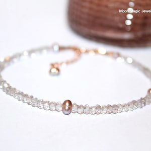 May include: A delicate bracelet with small, clear, faceted beads and three mauve-coloured pearls. The bracelet has a rose-gold clasp. The text "Moon Magic Jewellery" is visible in the background. The bracelet is displayed on a white surface.