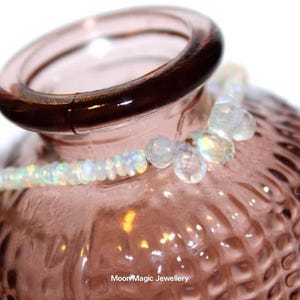 Puede incluir: Primer plano de un jarrón de cristal rosa con un borde marrón oscuro, adornado con un delicado collar de cuentas de ópalo y cristal. El collar está drapeado alrededor del jarrón, mostrando sus colores iridiscentes y su intrincado diseño. El jarrón tiene una superficie texturizada.