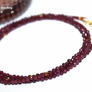 May include: A three-strand necklace composed of small, faceted, deep red beads. The necklace has gold-coloured clasps and accents. The text "Moon Magic Jewellery" is visible in the upper left corner.