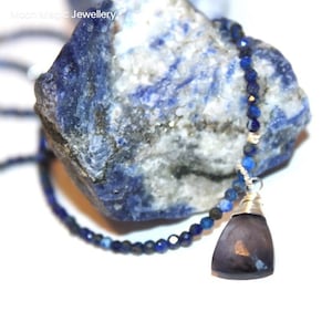 Op de afbeelding: Een zilveren ketting met een donkerpaarse edelsteen hanger en blauwe lapis lazuli kralen. De ketting wordt getoond op een blauw-witte steen.