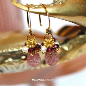 Könnte beinhalten: Ein Paar goldfarbene Ohrhänger. Jeder Ohrring hat eine Ansammlung kleiner, roter und weißer Perlen über einem rosa, tropfenförmigen Stein. Die Ohrringe hängen an goldfarbenen Haken. Der Text "Moon Magic Jewellery" befindet sich unten.
