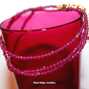 May include: A multi-strand necklace made of small, pink, faceted beads with gold-tone clasps. The necklace is wrapped around a deep red glass. The image is well-lit, showing the necklace's intricate details and the glass's rich colour. The text "Moon Magic Jewellery" is visible.