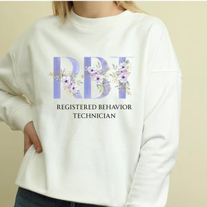 Op de afbeelding: Een witte sweatshirt met de letters "RBT" in lichtpaars, versierd met bloemmotieven. Daaronder staan de woorden "REGISTERED BEHAVIOR TECHNICIAN" in het zwart gedrukt. De sweatshirt heeft lange mouwen.
