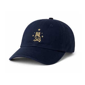 May include: Navy blue baseball cap with a curved brim. The cap features a gold embroidered owl design with stars. The cap is made of a soft fabric and has an adjustable strap at the back.