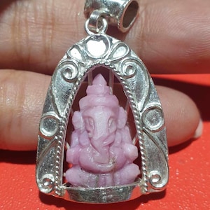 May include: A silver pendant featuring a carved pink gemstone of the Hindu deity Ganesha. The pendant is in the shape of an archway with intricate details.