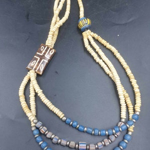 May include: A three-strand necklace with white beads, blue beads, and a brown wooden pendant with a geometric design. The necklace also features a blue and yellow bead.