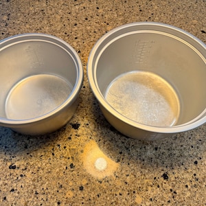 Może przedstawiać: Dwa srebrne garnki wewnętrzne do ryżowaru. Garnki są okrągłe z lekko rozszerzonym brzegiem. Na wewnętrznej stronie widoczne są oznaczenia pomiarowe. Garnki stoją na nakrapianym blacie.