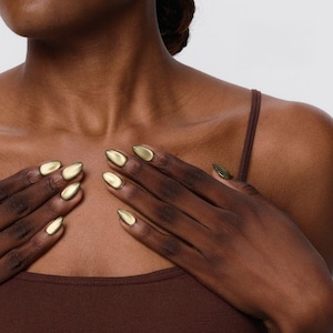 May include: Close-up of hands with gold-colored, almond-shaped artificial nails. The nails have a metallic finish and are a warm, golden hue. The person is wearing a brown tank top. The background is a plain, light color.