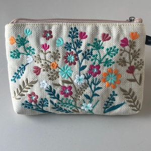 May include: A beige fabric cosmetic bag with a zipper closure and embroidered floral design in shades of pink, orange, green, and white. The bag has a small tag that reads "Made in China".