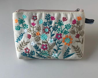 May include: A beige fabric cosmetic bag with a zipper closure and embroidered floral design in shades of pink, orange, green, and white. The bag has a small tag that reads "Made in China".