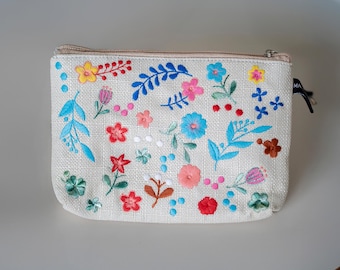 May include: A white canvas zippered pouch with a floral embroidery design. The embroidery features blue, pink, red, yellow, and green flowers and leaves.