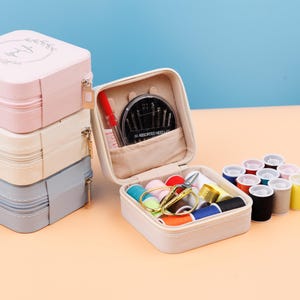 K&ouml;nnte beinhalten: Ein wei&szlig;es N&auml;hset mit Rei&szlig;verschluss. Das Set ist ge&ouml;ffnet und enth&auml;lt eine Vielzahl von N&auml;hzubeh&ouml;r, darunter Nadeln, Garn, einen Fingerhut und eine Schere. Das Set liegt auf einer hellbraunen Oberfl&auml;che.