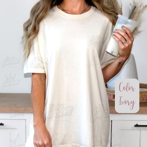 May include: Ivory-colored oversized t-shirt. The shirt has short sleeves and a relaxed fit. The text "Color Ivory" is displayed in a pink box. The model is wearing a necklace and earrings and holding a beverage.
