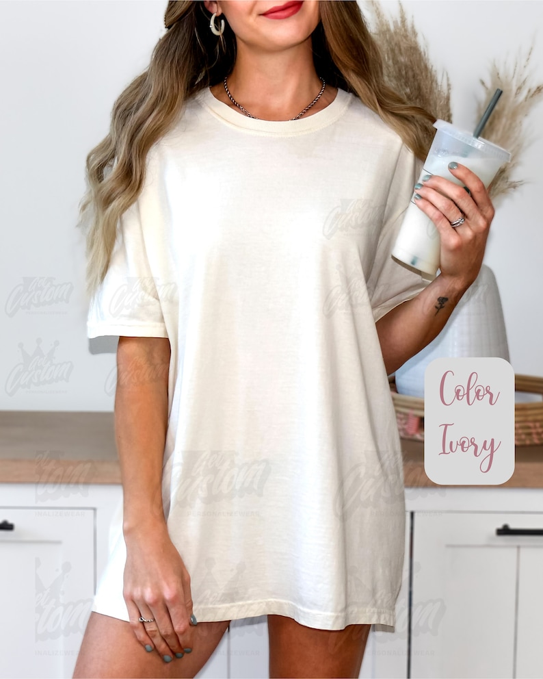 May include: Ivory-colored oversized t-shirt. The shirt has short sleeves and a relaxed fit. The text "Color Ivory" is displayed in a pink box. The model is wearing a necklace and earrings and holding a beverage.
