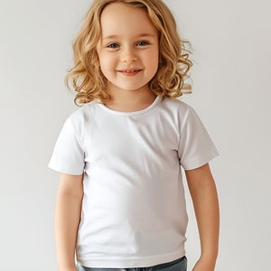 May include: A young girl with long, curly blonde hair smiles at the camera while wearing a white t-shirt.