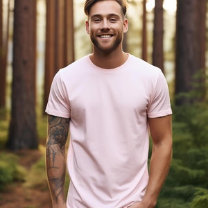 May include: A man with a light pink t-shirt and gray pants stands in a forest. He is smiling and has a tattoo on his left arm.