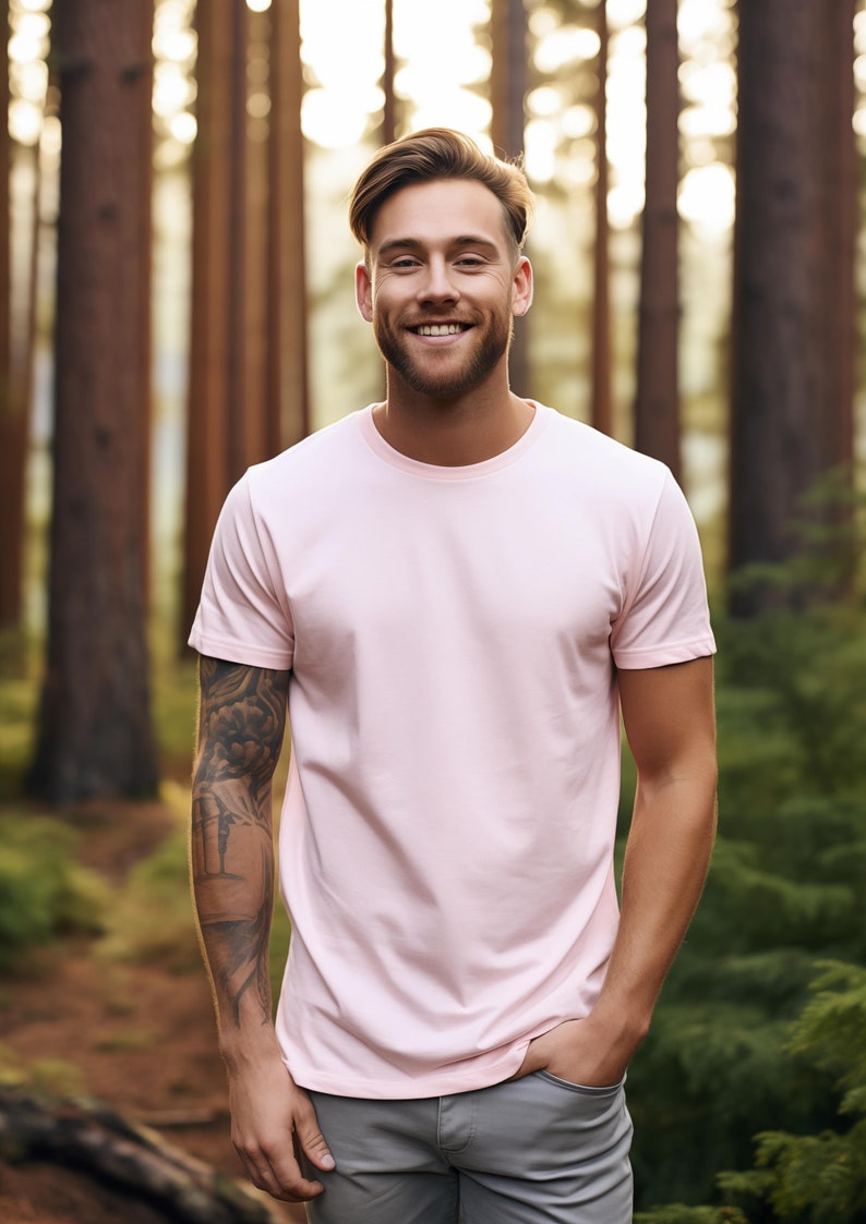 May include: A man with a light pink t-shirt and gray pants stands in a forest. He is smiling and has a tattoo on his left arm.