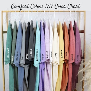 May include: A color chart of Comfort Colors 1717 t-shirts, showcasing a range of hues from light green to berry. The shirts are displayed on white hangers, with the color names printed below each shirt. The text "Comfort Colors 1717 Color Chart" is at the top.