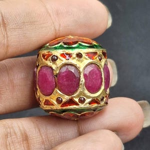 May include: A gold-colored, cylindrical bead with a vibrant design. The bead features oval-shaped, deep red gemstones set within a gold frame. The top and bottom sections are adorned with green, red, and orange enamel detailing, creating a striking contrast.