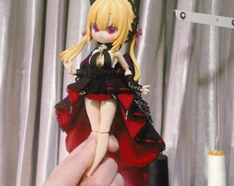 May include: A small doll with blonde hair and a red and black dress. The doll is wearing a crown and has red eyes. The doll is being held by a hand.