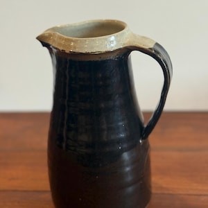Könnte beinhalten: Handgefertigter Keramikkrug mit schwarzem glasiertem Körper und hellbeigem Rand. Der Krug hat einen gebogenen Griff und einen Ausguss zum einfachen Ausgießen. Der Steinzeugkrug ist ein funktionaler und dekorativer Artikel zum Servieren von Getränken.