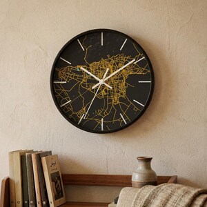 May include: A round wall clock with a black face featuring a detailed gold street map design. The clock has a thin black frame, white hour and minute hands, and white tick marks for each hour. The design displays a complex network of city streets.