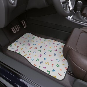 May include: White car floor mat with a colorful Arabic alphabet pattern. The mat is rectangular with a black border and is placed in the driver's side footwell of a car. The Arabic letters are in various colors.