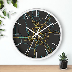 May include: A round wall clock with a white frame. The clock face features a detailed map design in yellow and teal against a black background. White hands indicate the time. The clock is on a wooden surface.