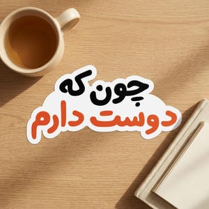 May include: A die-cut sticker featuring the Persian text "چون که دوست دارم" in black and orange lettering on a white background. The sticker is placed on a wooden surface next to a ceramic mug filled with tea and a closed notebook.