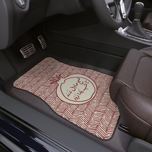 May include: Car floor mat with a red and cream chevron pattern and a cream-colored circle with text and a pomegranate illustration. The mat is in a car interior with a brown leather seat and black dashboard.